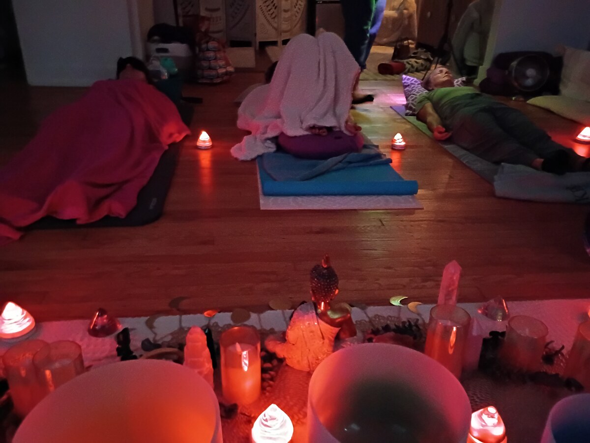 Crystal singing bowls, clear quartz points, pillar candles and a seated Buddha statue lit red and orange on the altar cloth at the Cosmic Virgo Recalibration sound bath in Ferndale, Michigan, with three guests resting on mats and blankets behind them