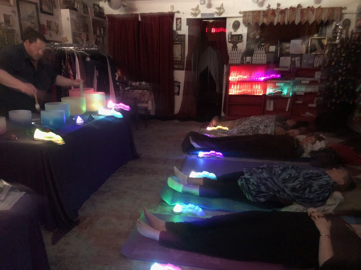 Rev. Michael strikes a frosted crystal singing bowl at a draped table while four people rest on mats under blankets in a dimly lit room