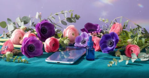 A phone resting face down on a teal linen cloth beside pink ranunculus, violet anemones, green eucalyptus and a small blue glass bottle in bright daylight