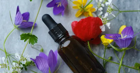 An amber dropper bottle of flower essence on gray stone, surrounded by purple bellflowers, a red poppy, yellow blossoms and small white wildflowers