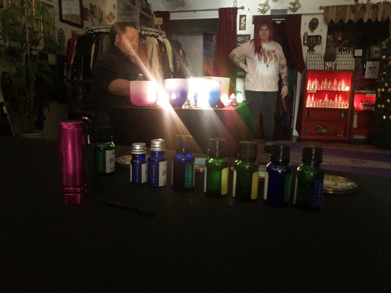 A row of small blue and green glass essential oil bottles along the front of a dark table, with a man playing three lit crystal singing bowls behind them and a woman standing beside red-lit retail shelves of bottles