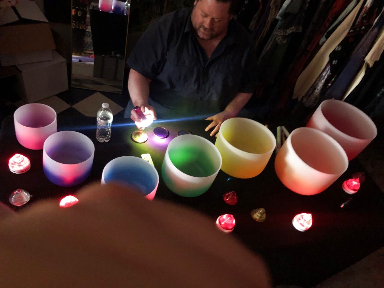 A man in a dark shirt seated behind seven frosted crystal singing bowls arranged in an arc on a black cloth, the bowls lit from inside in pink, white, blue, green, yellow and orange, with small red and pink tea lights scattered around them in a darkened room.