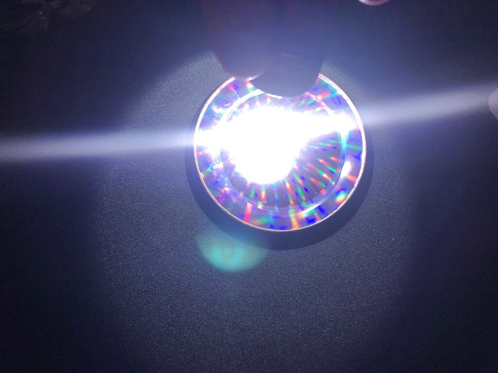 A round Pleiadian Cosmic Healing Disc lying on dark cloth with a bright light held behind it, splitting into a dense starburst of red, green and blue diffraction spokes